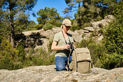 Tourist woman with cap and backpack taking a pair of binoculars to observe animals in the forest.