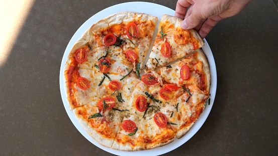 A person is grabbing a slice of California-style pizza from a white plate, showcasing the delicious combination of ingredients, fast food perfection.
