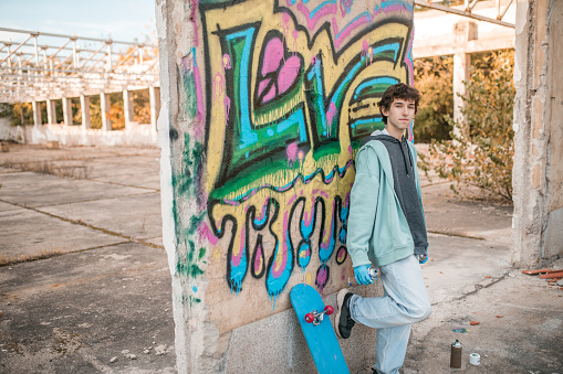 Street artist holding sprays and standing leaning against the wall of an abandoned building on which he has drawn graffiti