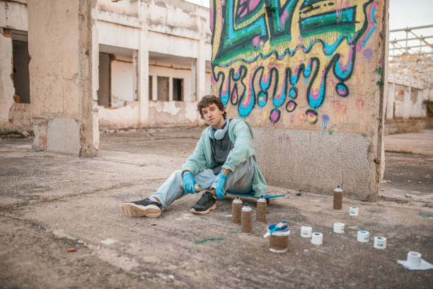 Portrait of a talented young street artist with headphones around his neck sitting in front of the wall of an abandoned building, on which he has drawn beautiful, colorful graffiti
