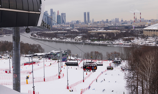Skiers are being transported uphill on a ski lift at a snowy resort in Moscow, Russia, with a river and buildings in the background