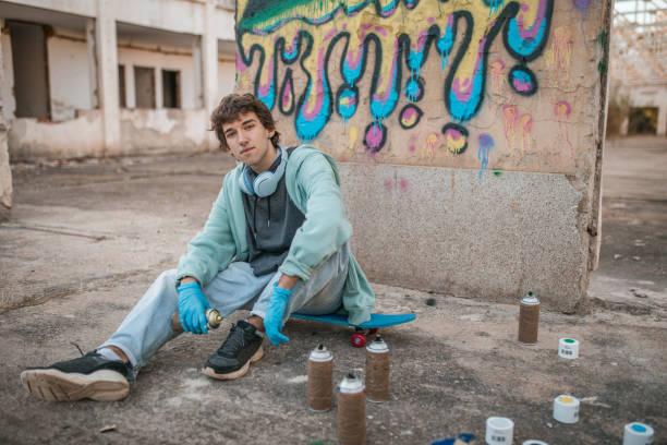 Portrait of a talented young street artist with headphones around his neck sitting in front of the wall of an abandoned building, on which he has drawn beautiful, colorful graffiti