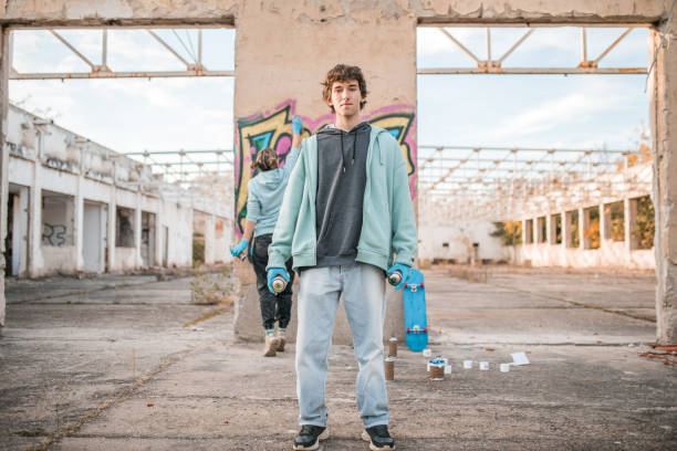 Portrait of a young artist looking directly into the camera while holding a graffiti spray, with his friend drawing graffiti in the background