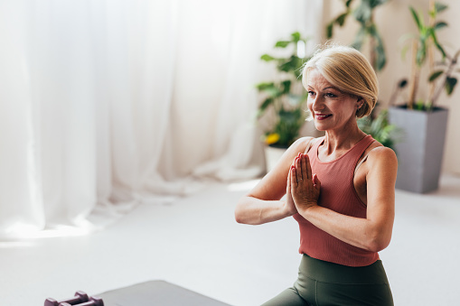 A serene senior woman in workout attire demonstrates a yoga pose in a well-lit, peaceful room, promoting health and mindfulness.