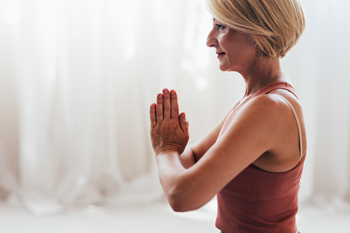 Side view of a serene mature woman practicing yoga with hands in prayer position, conveying tranquility and wellness at home.