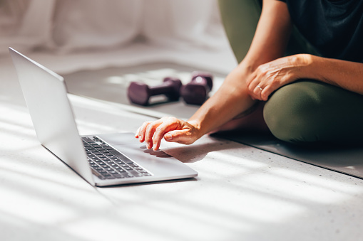 An active woman relaxes on the floor, pausing her workout to interact with a laptop, with dumbbells nearby, in a serene home environment.