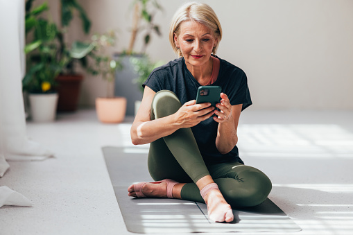 Active senior woman sitting on a yoga mat, using her smartphone in a bright, plant-filled room, enjoying a healthy lifestyle.