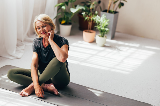 A content senior woman enjoying a yoga session inside a bright, plant-filled room, radiating positivity and well-being.