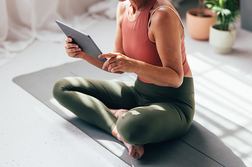 Fit female in activewear seated on a yoga mat, browsing a tablet in a sunlit room with plants in the background.
