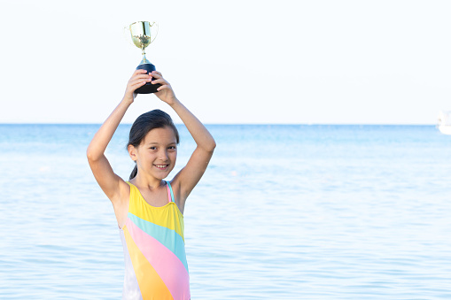 Little girl wearing swimwear  is holding golden trophy award with a toothy smile and is looking at camera and is standing in sea. Representing swimming contest winner and first price of competition.