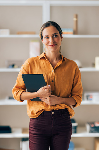 A smiling woman in casual office attire confidently holds a tablet, exuding friendliness and competence in a contemporary workspace.