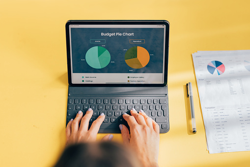 Overhead view of a professional working on a tablet analyzing a colorful budget pie chart. Financial documents scattered on a yellow background.