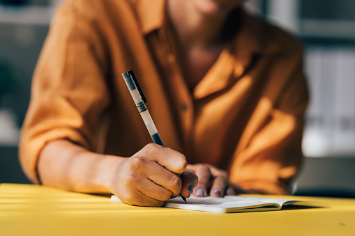 Close-up of a person's hand writing in a notebook placed on a vibrant yellow desk, depicting concentration and work.