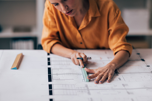 Focused woman architect working on construction blueprints at office desk, using ruler and pen, planning.