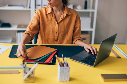 A focused professional examines fabric samples while working on a design project at a vibrant yellow desk with a laptop.