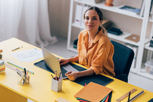 Confident female professional works on a laptop at a vibrant yellow desk in a modern home office setting.