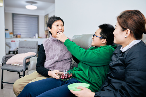 Happy Chinese multi-generational family sitting on the sofa eating fruit together, the playful grandson puts a cherry into the grandmother's mouth