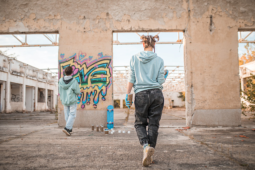 Back view of two friends drawing graffiti on the wall of an abandoned building