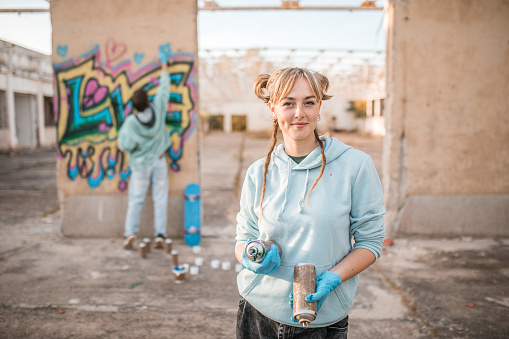 Portrait of a young artist looking directly into the camera while holding a graffiti spray, with her friend drawing graffiti in the background