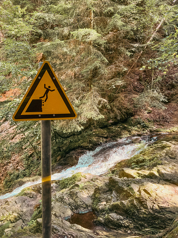 Yellow sign warning hikers of the risk of falling off of a steep mountain cliff. Hazardous area on a tall cliff above a mountain stream. Located within the Black Forest in Germany. Breitnau, Breisgau-Hochschwarzwald, Baden-Württemberg, Germany.