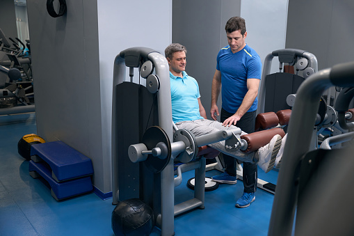 Pleased athletic guy performing seated leg extension on exercise machine supervised by fitness trainer