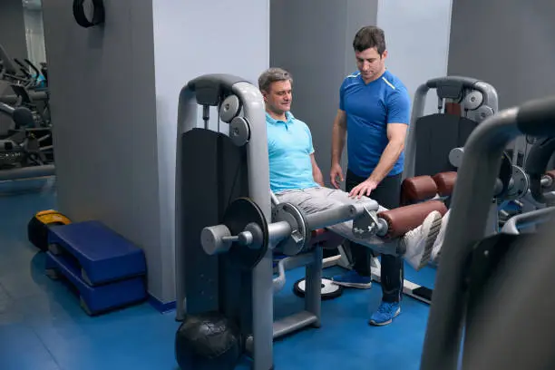 Pleased athletic guy performing seated leg extension on exercise machine supervised by fitness trainer