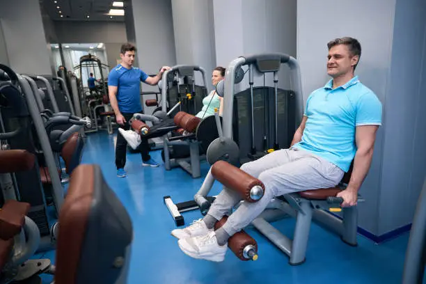 Smiling woman and man performing seated leg extension on gym equipment under guidance of trainer