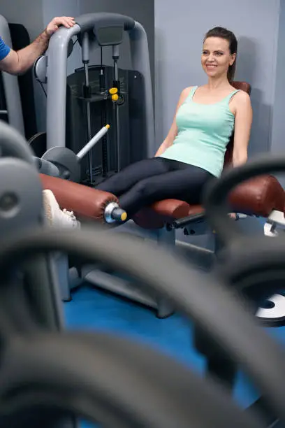 Pleased athletic female doing seated leg extension on fitness equipment in presence of personal trainer