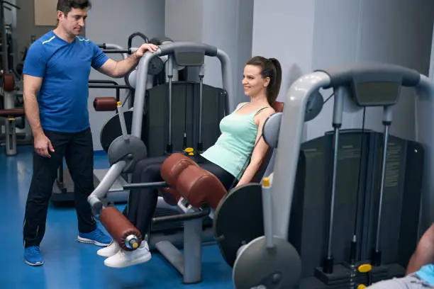 Cheerful mature lady performing seated leg extension on exercise machine under guidance of gym instructor