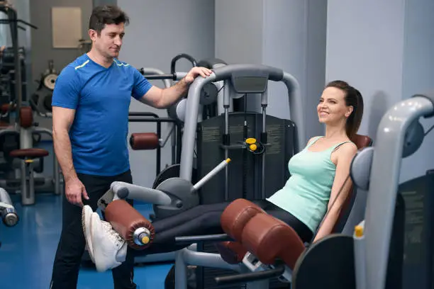 Smiling sporty woman performing seated leg extension on gym equipment under guidance of personal trainer
