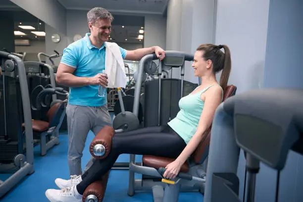 Cheerful fit lady working out on leg extension machine while talking to joyous fitness partner