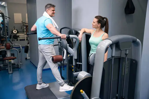 Pleased sporty man performing side leg raise on exercise machine in presence of smiling fitness partner