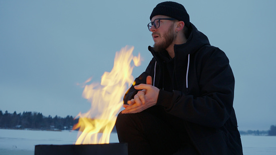 Man warms hands by fire on frozen lake at dusk