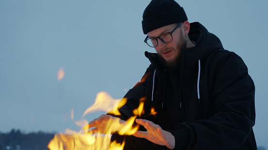 Man warms hands by fire on frozen lake at dusk