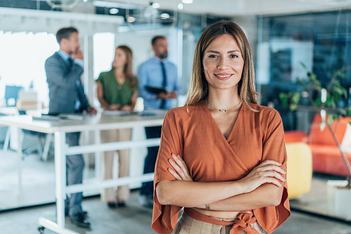 Portrait of a young business woman in the modern office, and a team working behind her. Successful businesswoman standing in creative office and looking at camera.