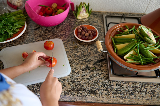 Female chef slicing tomatoes, standing by kitchen counter, preparing ingredients for cooking a delicious authentic vegetarian tagine, according to traditional Moroccan recipe in the kitchen at home