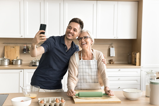 Cheerful adult son and mature mom taking selfie on smartphone while baking in kitchen, rolling dough, talking on video call, looking at mobile phone having fun, enjoying family communication