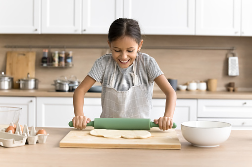 Happy preteen baker child girl in chef apron baking alone in home kitchen, rolling dough for pitta on table with bakery food ingredients, preparing homemade pastry dessert, enjoying culinary hobby