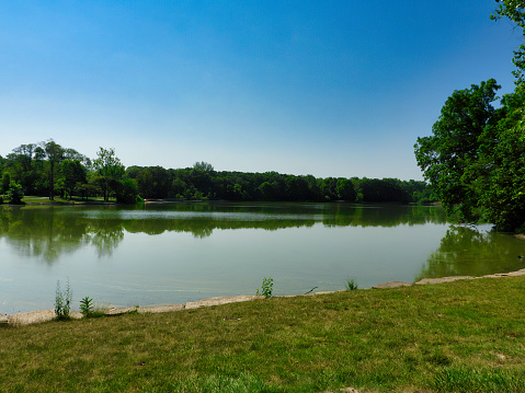 Lake at Park on a Sunny Summer Day with Bright Green Trees Along Lakeshore and Reflected in Calm Water with Bright Blue Sky in Background