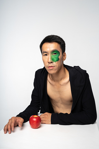 Asian man with green face paint and apple on table in a conceptual portrait