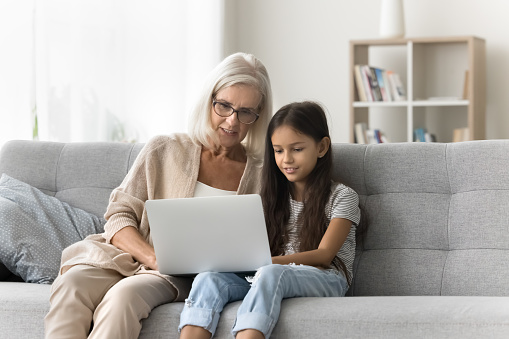 Focused grandmother helping preteen granddaughter kid with school homework task, talking to girl using laptop computer on home couch. Grandma and kid resting with online gadget on sofa