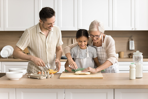 Happy kid, dad and grandma baking in home kitchen together, rolling dough, beating up eggs for breakfast, preparing tasty homemade bakery dessert, pastry food for family dinner