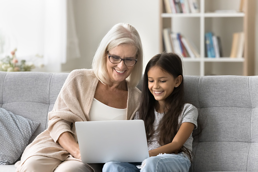 Happy grandma and pre teen kid girl using online learning application on laptop together, sitting on home sofa, holding computer on laptop, smiling, laughing, enjoying leisure, watching movie