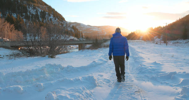 Mature man hikes towards sunrise in snowy winter landscape with snowcapped mountains distant