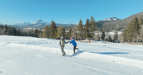 Mature man offers helping hand to woman on hike in snowy winter landscape with snowcapped mountains distant