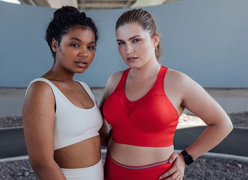 Portrait of two young plus-size females in sportswear. Two women of different ethnicities standing together after workout outdoors.