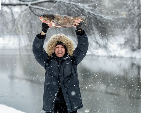 A fisherman on winter fishing caught a large silver carp and holds it in his hands. He rejoices at the good catch, despite the cold and snowfall.