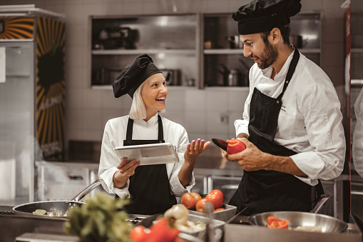 Modern Kitchen Restaurant: Male and Female Chefs Uses Digital Tablet Computer While Working in a Modern Professional Kitchen. Preparing Gourmet Organic Dishes