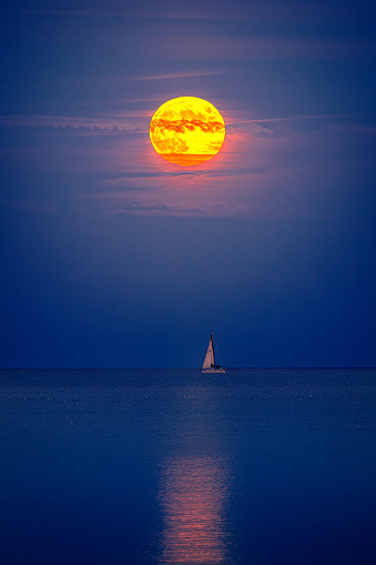 View of a colourful full moon over Lake Ontario with a sailboat on the horizon.