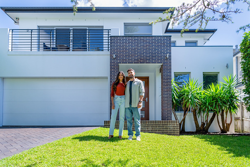 Couple standing proudly in front of their new home. They are both wearing casual clothes and embracing. They are looking at the camera and smiling and he is holding a key to the house. The house is contemporary with a brick facade, driveway, balcony and a green lawn. The front door is also visible. Copy space